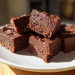 Close-up of rich, dark chocolate fudge squares stacked on a white plate, highlighting the creamy texture of the fudge.