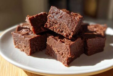 Close-up of rich, dark chocolate fudge squares stacked on a white plate, highlighting the creamy texture of the fudge.