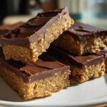 A stack of homemade, no-bake protein bars with a thick layer of chocolate topping resting on a white plate.