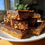A stack of homemade chocolate-topped protein bars with visible nuts and seeds on a white plate.
