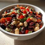 A white bowl filled with colorful three-bean salad featuring kidney beans, white beans, red peppers, and herbs.