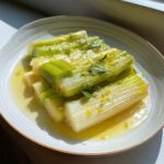 A plate of tender braised leeks swimming in a light sauce, topped with fresh herbs and lemon zest.
