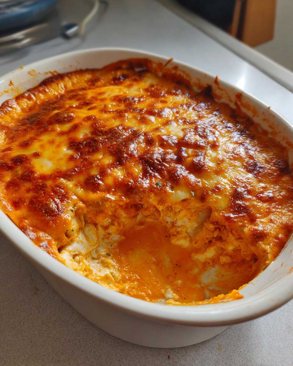 A close-up of hot, baked buffalo chicken dip in a white casserole dish with a scoop removed, showing the creamy orange sauce and melted cheese.
