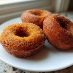 Close-up of three freshly baked pumpkin donuts coated in cinnamon sugar resting on a white plate.
