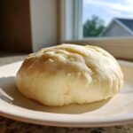 A smooth, pale yellow ball of homemade marzipan recipe dough resting on a white plate near a window.