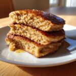 Close-up of a stack of three thick banana oatmeal pancakes drizzled with syrup on a white plate.