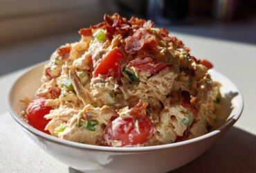 A close-up of creamy blt chicken salad mixed with tomatoes and topped generously with crispy bacon bits in a white bowl.