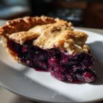 A close-up of a generous slice of blueberry pie recipe showing juicy filling and a golden, flaky crust.