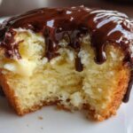 A close-up of a boston cream pie cupcake with a bite taken out, showing vanilla cream filling and rich chocolate glaze dripping down.