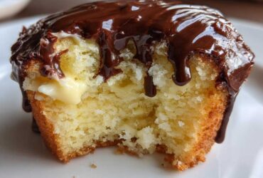 A close-up of a boston cream pie cupcake with a bite taken out, showing vanilla cream filling and rich chocolate glaze dripping down.