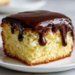 A close-up of a square slice of cake resembling a boston cream pie cupcake, topped with dripping dark chocolate ganache and visible vanilla cream filling.