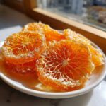 Close-up of bright orange, crystallized candied orange slices piled on a small white plate near a window.