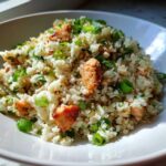 A close-up of a white bowl filled with cauliflower fried rice mixed with pieces of browned chicken and topped with fresh green onions.