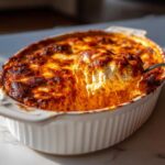 A spoonful of hot, cheesy buffalo chicken dip being lifted from a white baking dish with a browned top.