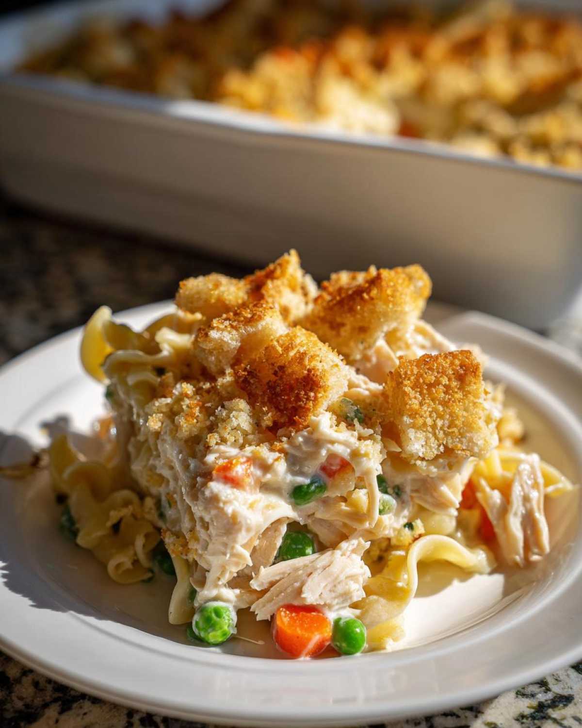 A serving of creamy chicken noodle casserole with visible chicken, peas, carrots, and a crunchy breadcrumb topping.