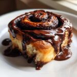 Close-up of a fluffy chocolate cinnamon roll generously covered in thick, glossy chocolate glaze.