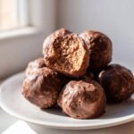 A stack of rich, chocolate-coated coconut balls, with one bitten open to show the texture inside.