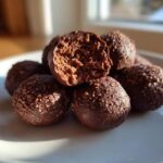 A pile of rich chocolate coconut balls on a white plate, with one bitten open revealing the textured interior.