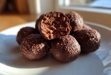 A pile of rich chocolate coconut balls on a white plate, with one bitten open revealing the textured interior.