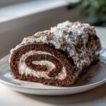A close-up of a rich chocolate yule log cake slice showing the dark sponge rolled with white cream filling, covered in ganache and powdered sugar.