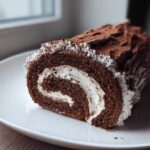 A cross-section slice of a rich chocolate yule log cake filled with white cream, dusted with cocoa powder.