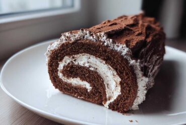 A cross-section slice of a rich chocolate yule log cake filled with white cream, dusted with cocoa powder.