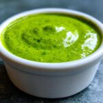 Close-up of vibrant green cilantro lime sauce served in a small white dipping bowl on a dark surface.