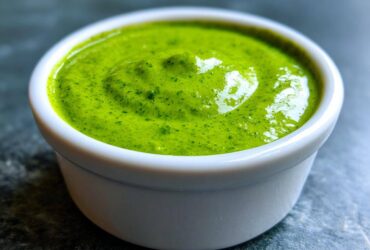 Close-up of vibrant green cilantro lime sauce served in a small white dipping bowl on a dark surface.