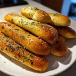 A close-up of several golden-brown copycat breadsticks seasoned with salt and green herbs on a white plate.