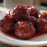 A close-up shot of several glistening cranberry meatballs coated in a thick, dark red glaze, served on a white plate.