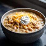 Close-up of a hearty bowl of creamy chicken chili topped with shredded cheese and a dollop of sour cream.