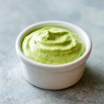 A close-up of thick, vibrant green cilantro lime sauce swirled in a small white dipping bowl.