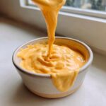 A thick stream of vibrant orange nacho cheese sauce is being poured into a small ceramic bowl, showing off its creamy texture.