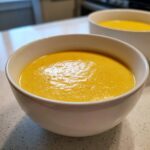 A close-up view of a creamy, vibrant orange cheese soup served in a white bowl on a speckled countertop.