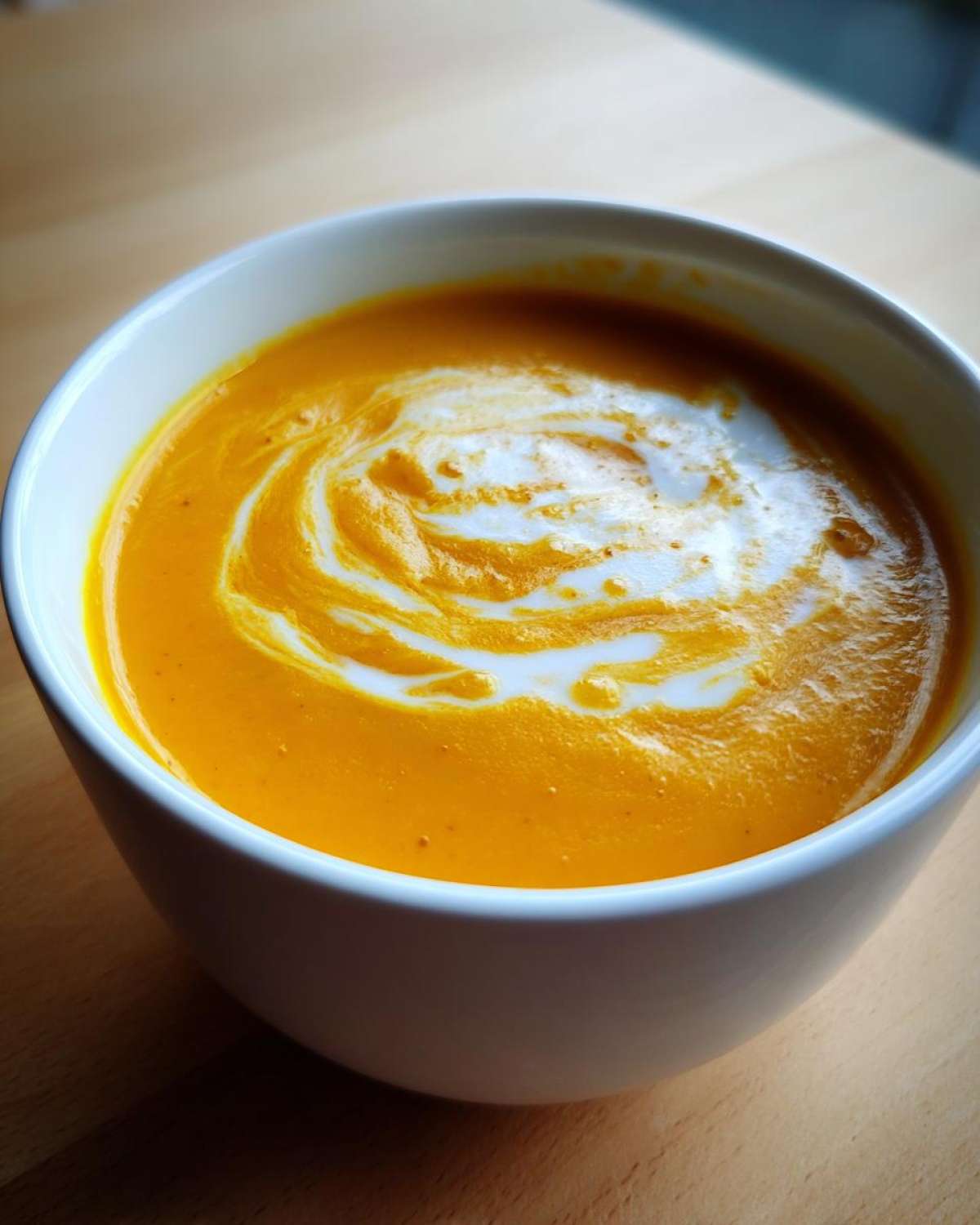 Close-up of vibrant orange squash soup garnished with a swirl of cream in a white bowl.