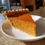 A perfect slice of creamy sweet potato pie with a crimped crust, sitting on a white plate in natural sunlight.