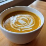 Close-up of a bowl of vibrant orange squash soup topped with a decorative swirl of cream and black pepper.