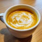 Close-up of vibrant orange squash soup in a white mug, topped with a swirl of cream.