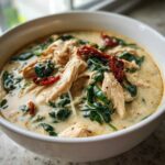 Close-up of a white bowl filled with creamy tuscan chicken soup, featuring shredded chicken, wilted spinach, and sun-dried tomatoes.