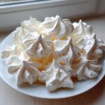 A close-up of freshly piped, crisp meringue cookies piled high on a white plate.
