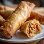 Close-up of several golden brown, crispy fried egg rolls, one cut open showing the savory filling.