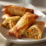 Close-up of several golden brown, crispy egg roll appetizers, one cut open to show the vegetable and meat filling.