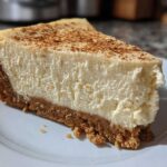 A close-up of a thick slice of creamy eggnog cheesecake with a graham cracker crust, dusted with cinnamon.