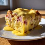 A square serving of rich eggs benedict casserole topped with dripping hollandaise sauce on a white plate.