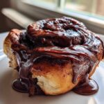 A close-up of a single, gooey chocolate cinnamon rolls smothered in thick, melted chocolate frosting.