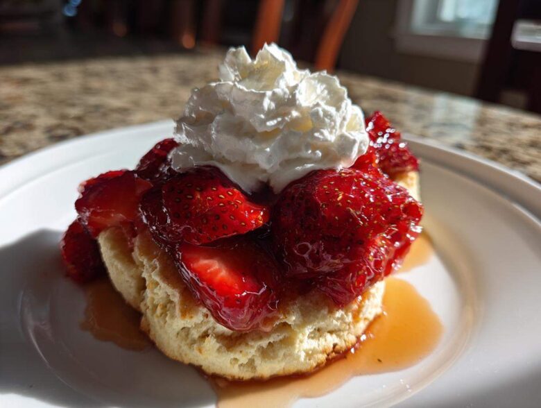 Close-up of a flaky shortcake topped with macerated strawberries and a dollop of whipped cream.