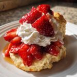 A close-up of a flaky shortcake topped with macerated strawberries and a generous dollop of whipped cream.