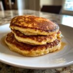 A close-up of a stack of three golden-brown buttermilk pancakes drizzled with syrup on a white plate.