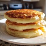A close-up of three thick, fluffy buttermilk pancakes stacked on a white plate, golden brown from cooking.