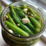 Close-up overhead view of bright green dilly beans being pickled with garlic cloves and dill in a glass jar.